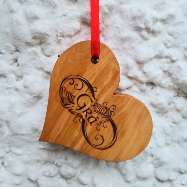 Wooden love heart decoration engraved with Gaelic word for "Love" - "Grá". With red ribbon hanging on a white wall.