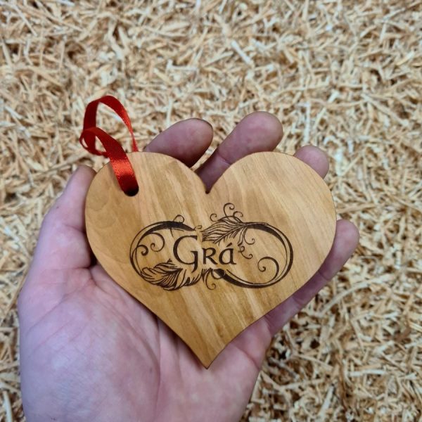 A hand holding a wooden love heart decoration engraved with Gaelic word for "Love" - "Grá". With red ribbon attached.